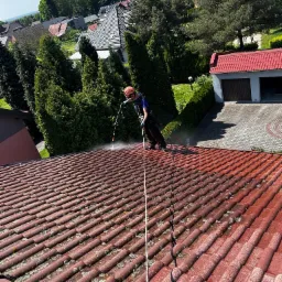 Pracownik w uprzęży alpinistycznej myje dach z czerwonej dachówki za pomocą lancy ciśnieniowej, widoczne zabrudzenia i mech na dachówce oraz budynki w tle.