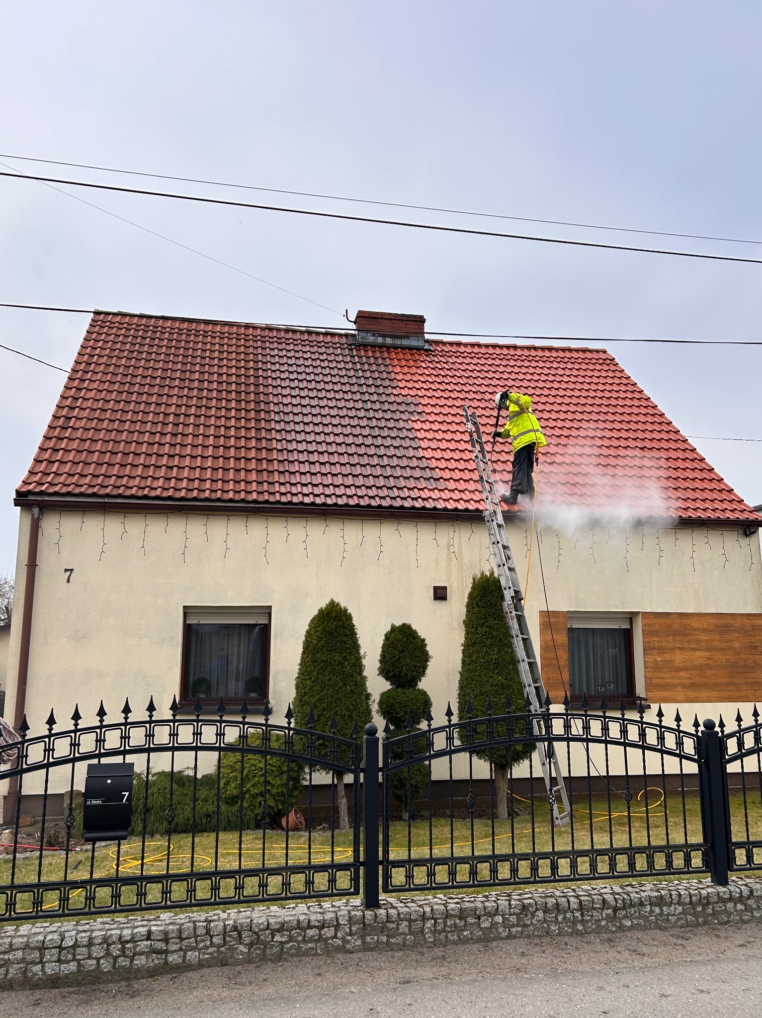 Pracownik w żółtej kurtce na drabinie czyści dach z czerwonej dachówki za pomocą myjki ciśnieniowej; widoczna różnica w czystości połowy dachu.