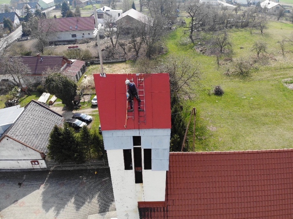 Pracownik na drabinie, na czerwonym dachu wieży kościoła, w trakcie prac konserwacyjnych, widok z góry na wieś.