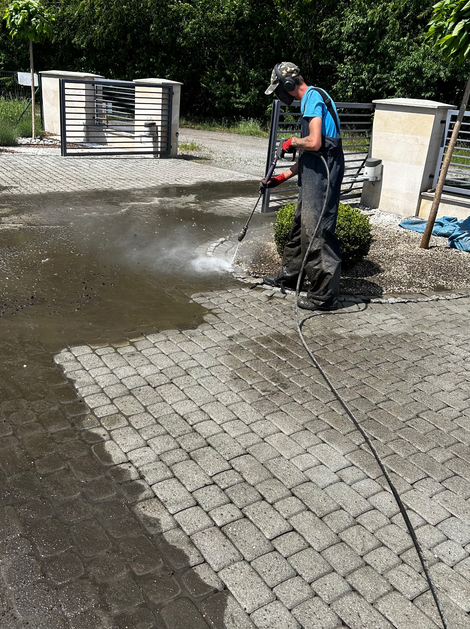 Pracownik w kombinezonie i słuchawkach ochronnych czyści kostkę brukową myjką ciśnieniową, widoczna różnica między umytą a brudną powierzchnią, w tle brama wjazdowa.