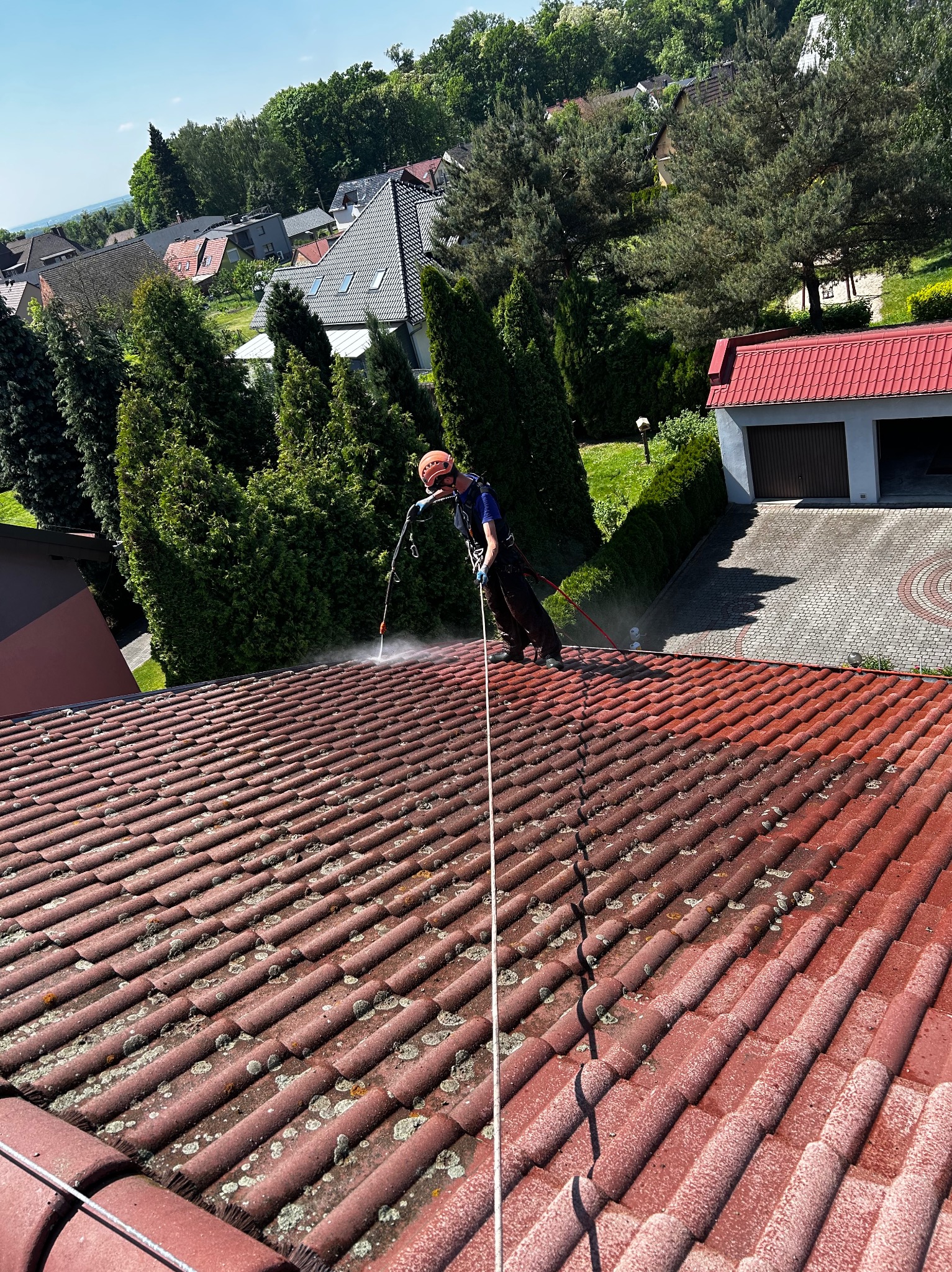 Pracownik w uprzęży alpinistycznej myje dach z czerwonej dachówki za pomocą lancy ciśnieniowej, widoczne zabrudzenia i mech na dachówce oraz budynki w tle.