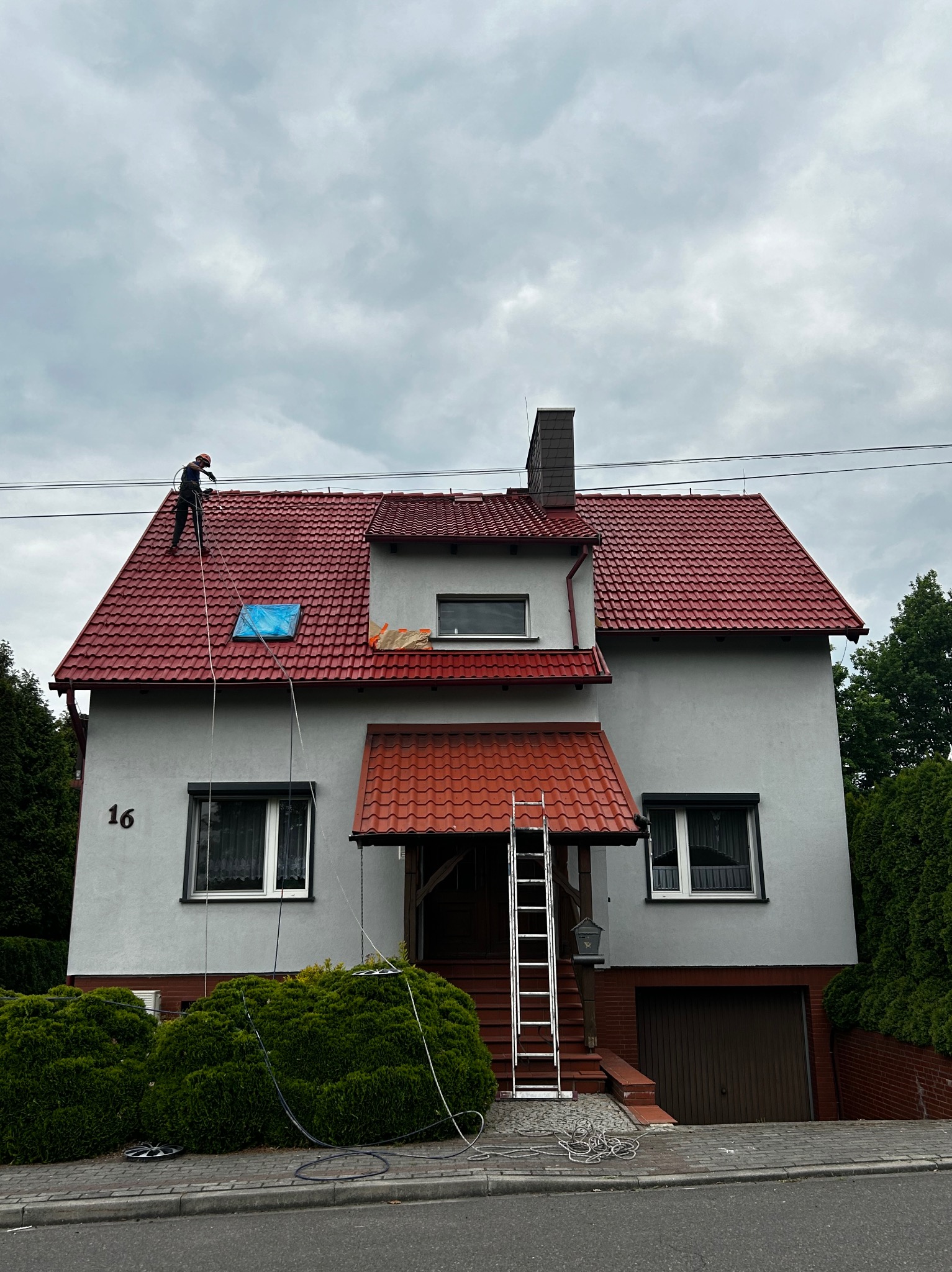 Pracownik w kombinezonie i kasku czyści czerwoną dachówkę dwupiętrowego domu z numerem 16, używając myjki ciśnieniowej; na dachu widoczne niebieskie zabezpieczenie świetlika.