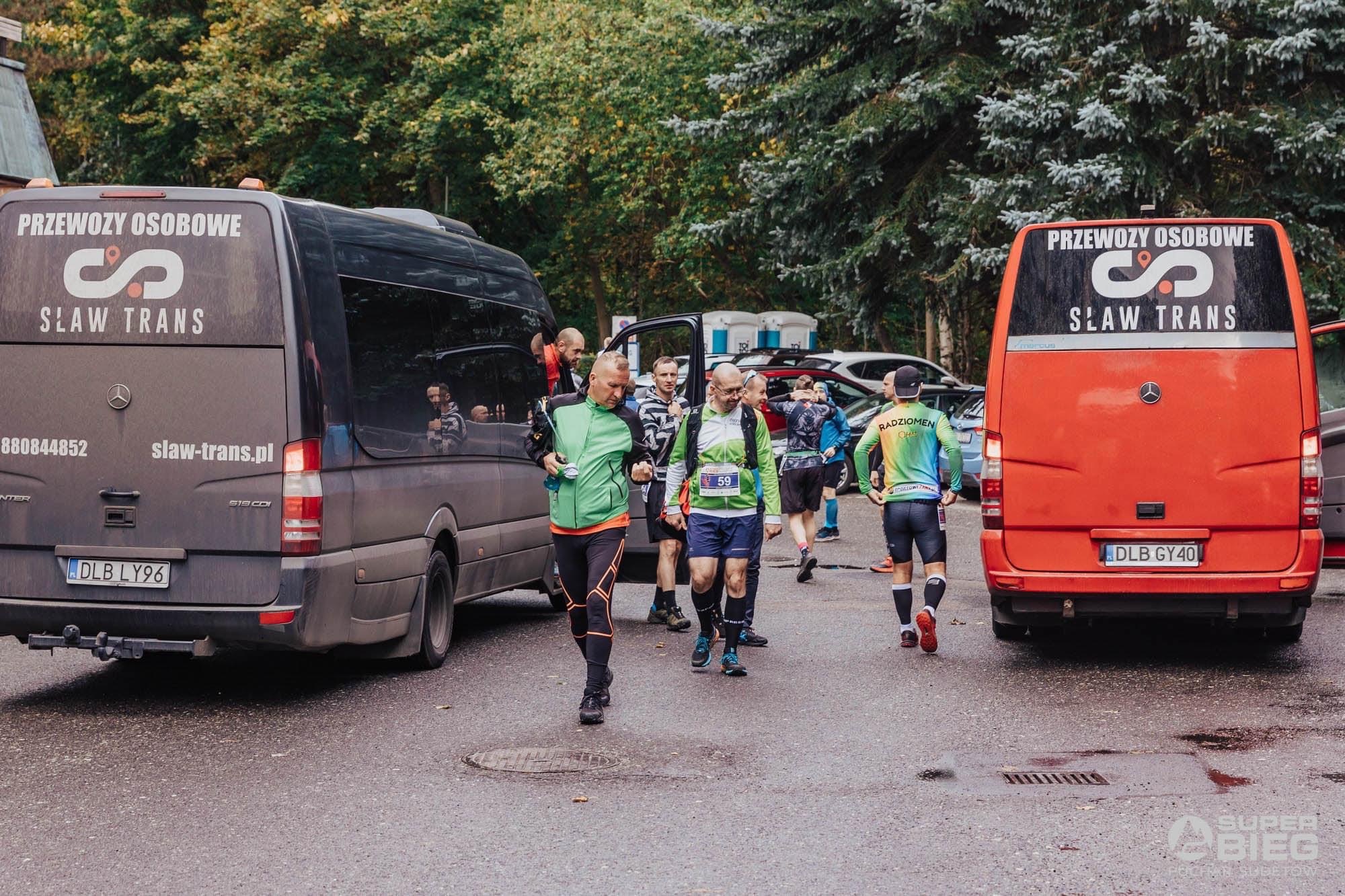 Dwa busy firmy Sław Trans z napisem 'Przewozy Osobowe' na tle grupy biegaczy po zawodach, z widocznymi numerami startowymi i logiem 'Super Bieg'.