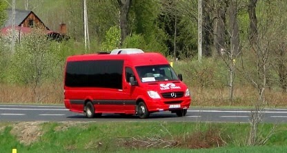 Czerwony bus Mercedes-Benz Sprinter z przyciemnianymi szybami, logo firmy na masce, jadący po asfaltowej drodze na tle zieleni.