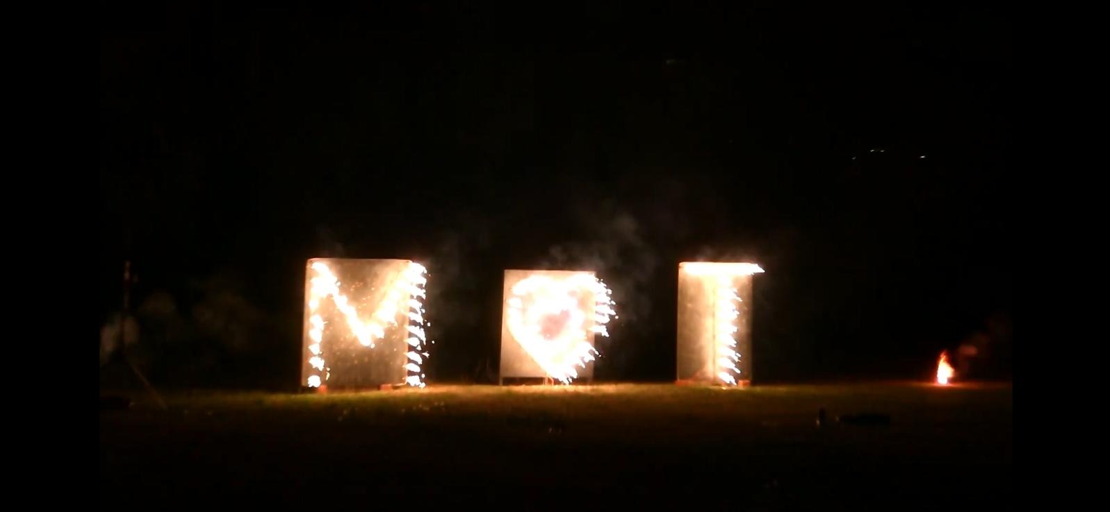 Nocny pokaz pirotechniczny: płonące litery 'MOT' na metalowych konstrukcjach, iskry i dym w ciemności. Unikalne oświetlenie i widowiskowy efekt specjalny.