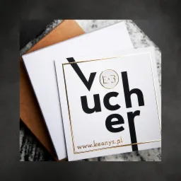 Voucher prezentowy na sesje fotograficzne z złotym obramowaniem i adresem strony internetowej, umieszczony na pudełku i marmurowym tle.