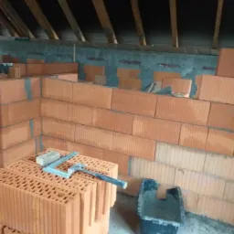 Wznoszenie ściany z czerwonych, dziurawych cegieł na poddaszu, widoczna czarna kuweta i poziomica na cegłach.