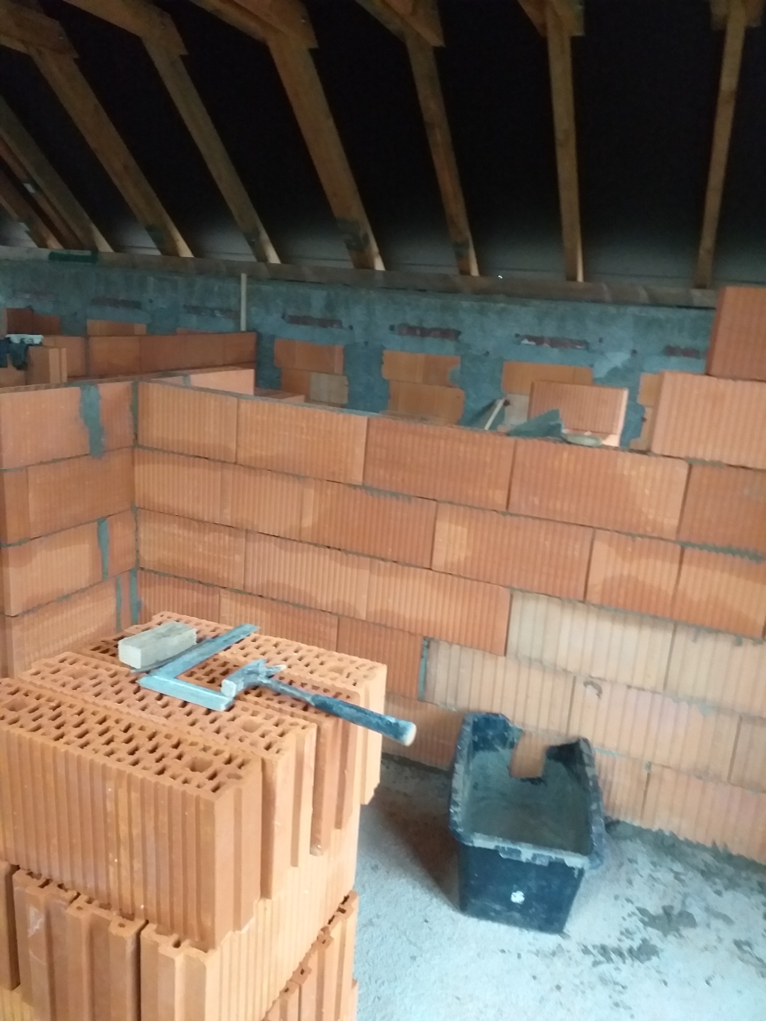 Wznoszenie ściany z czerwonych, dziurawych cegieł na poddaszu, widoczna czarna kuweta i poziomica na cegłach.