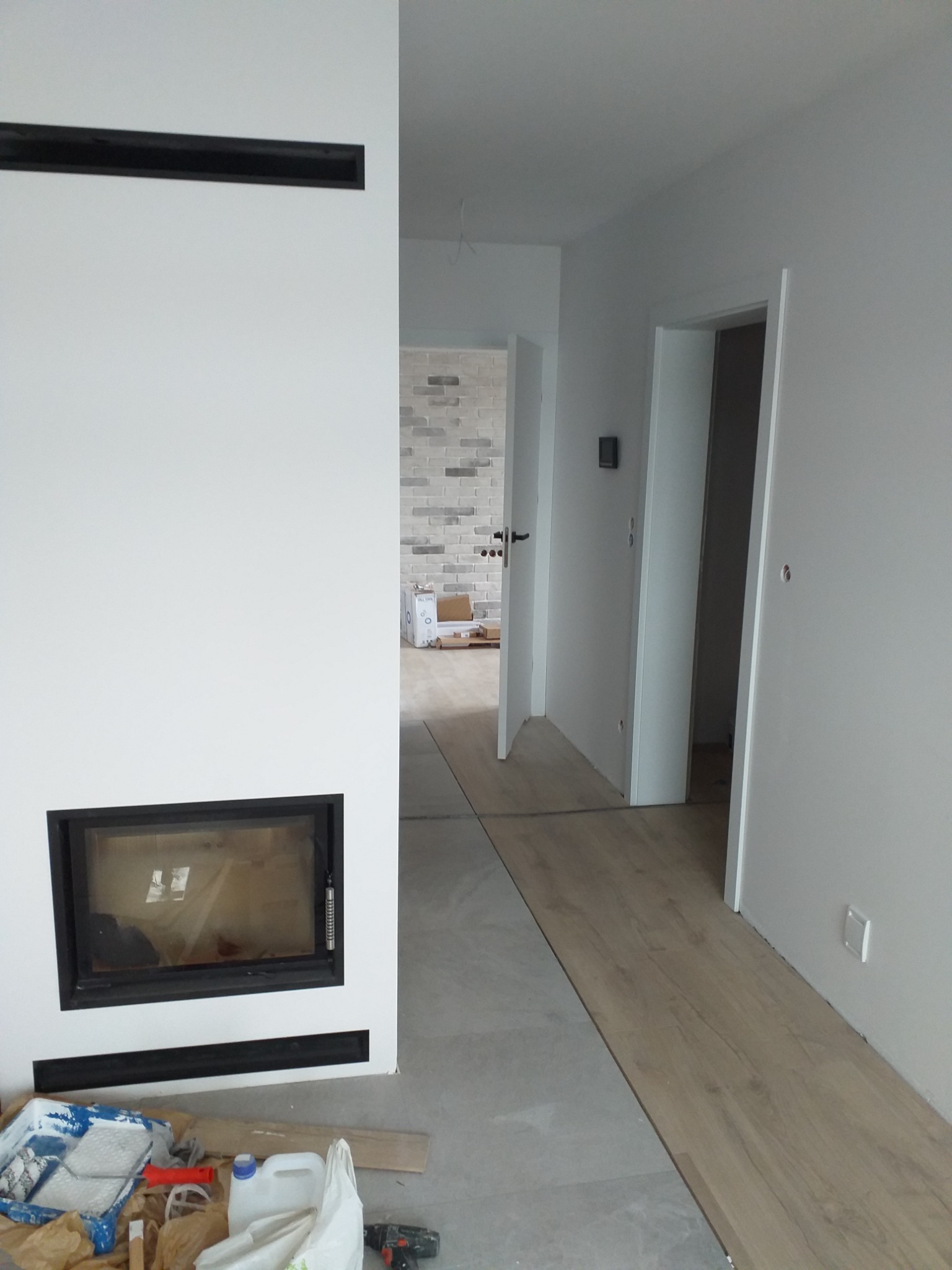 Wnętrze w trakcie wykańczania: biała ściana z czarnym obramowaniem kominka, podłoga częściowo wyłożona szarą płytką i panelami, widoczne narzędzia malarskie i wiertarka.