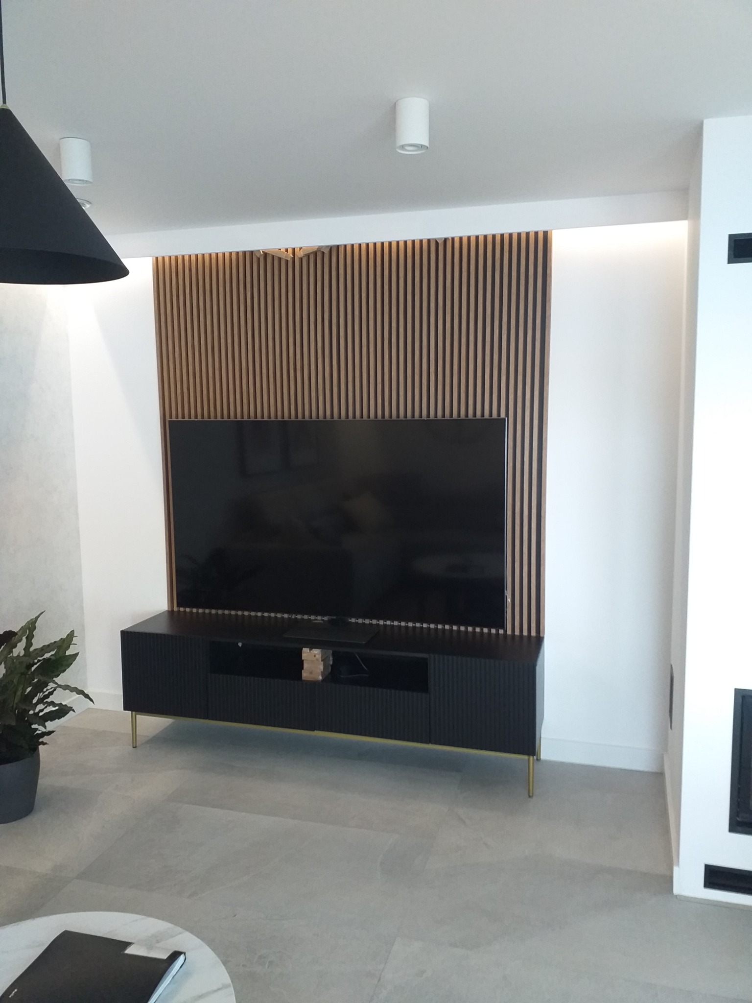 Nowoczesny salon z telewizorem na tle drewnianej ściany z lameli, czarna szafka RTV na złotych nóżkach, szara podłoga i fragment okrągłego stolika kawowego z książką.