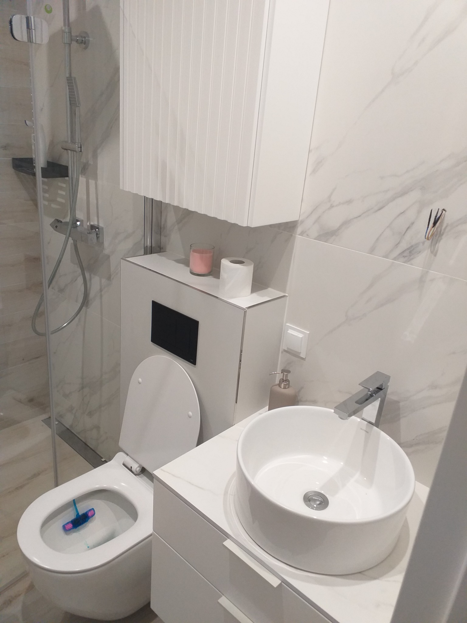 Nowoczesna, biała łazienka z marmurowymi płytkami, okrągłą umywalką nablatową, podwieszaną toaletą z czarnym panelem sterującym i szklaną kabiną prysznicową.