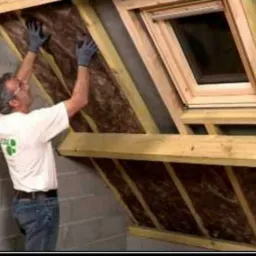 Pracownik w rękawicach montuje izolację z wełny mineralnej pod skośnym dachem z oknem dachowym, widoczna konstrukcja drewniana i ściana z bloczków betonowych.