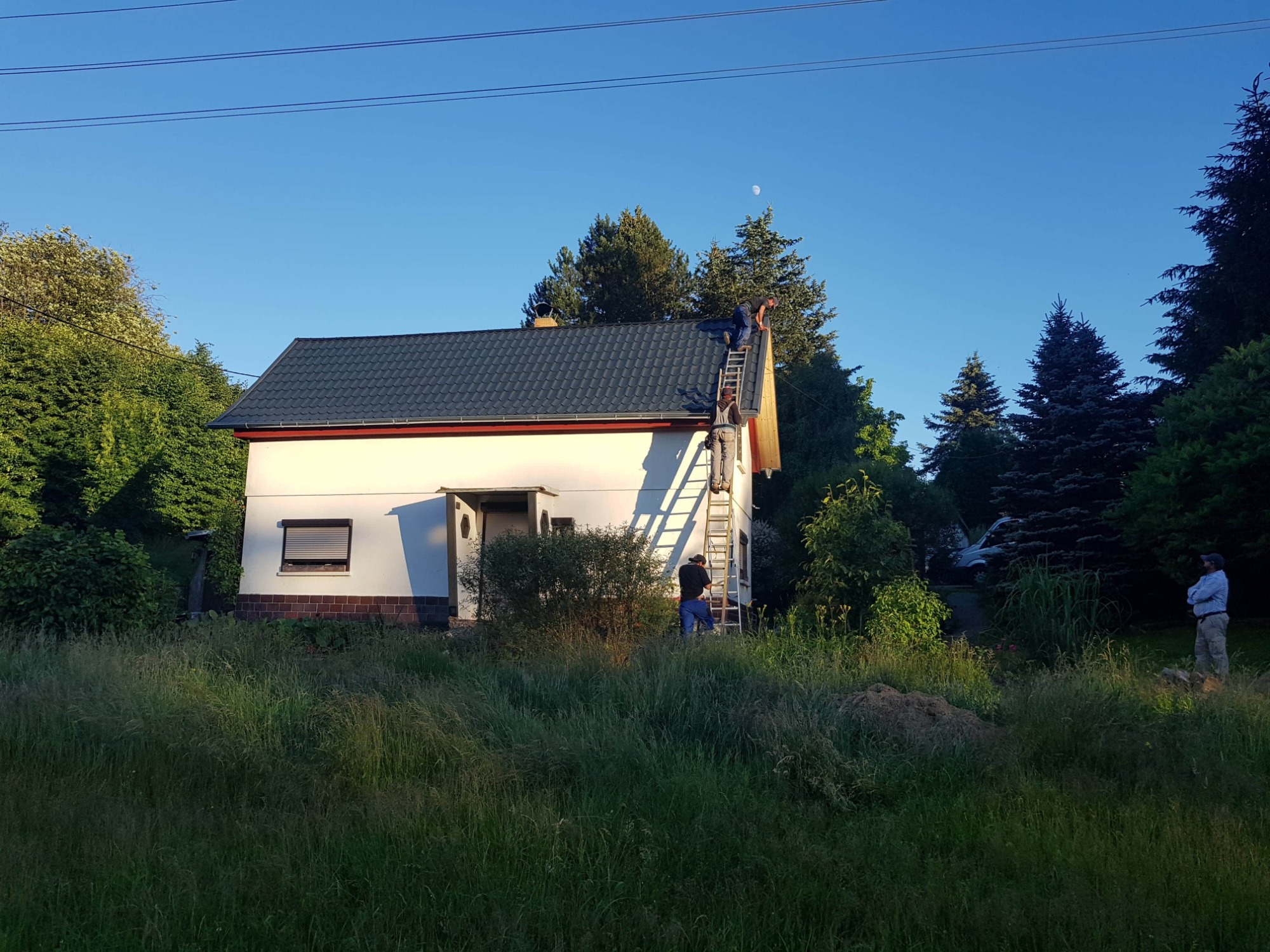 Dwóch robotników na drabinie przy dachu domu jednorodzinnego, jeden na dachu, drugi na drabinie, widoczne fragmenty nowej konstrukcji dachu, w tle błękitne niebo i księżyc.