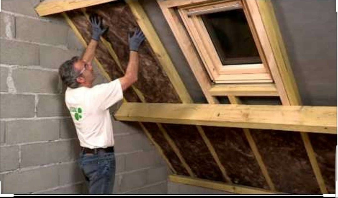 Pracownik w rękawicach montuje izolację z wełny mineralnej pod skośnym dachem z oknem dachowym, widoczna konstrukcja drewniana i ściana z bloczków betonowych.