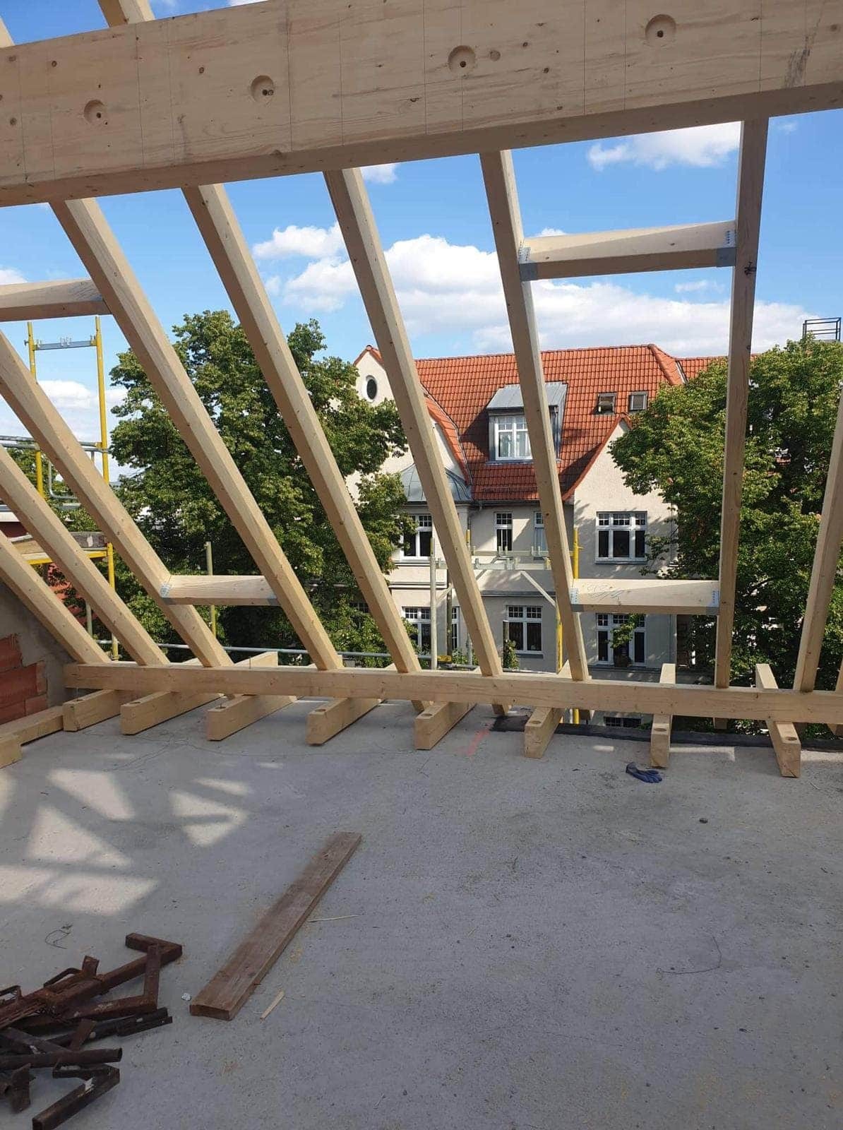 Wznoszenie drewnianej więźby dachowej z widokiem na sąsiedni budynek z czerwonym dachem i zielonymi drzewami w tle.