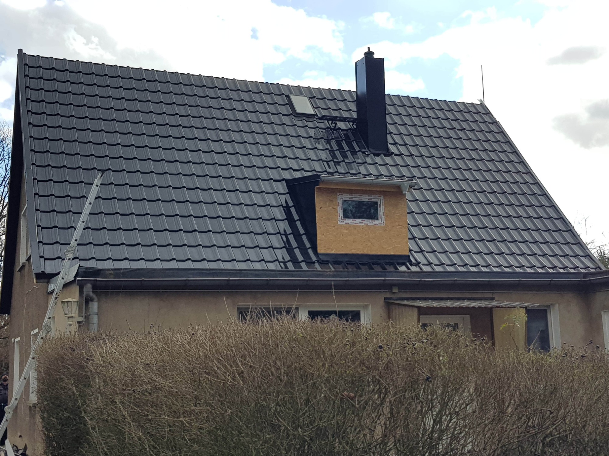 Dom z czarną dachówką karpiówką, nowym oknem dachowym w surowej obudowie z płyty OSB, czarnym kominem, drabina oparta o rynnę.