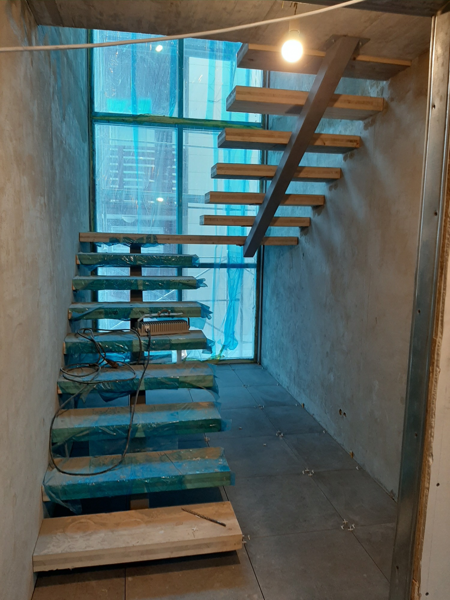 Montaż drewnianych schodów dywanowych z metalową konstrukcją nośną w surowym wnętrzu. Stopnie zabezpieczone folią ochronną, widoczne okno w tle, surowe ściany i ułożona podłoga z płytek.