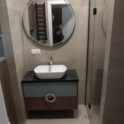 Perfect Home Rafał Potrawka - Nowoczesna łazienka z okrągłym lustrem, białą umywalką na szafce w odcieniach szarości i brązu oraz kabiną prysznicową z przezroczystego szkła.