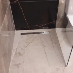 Nowoczesna łazienka z beżowymi płytkami imitującymi marmur, czarną ścianą z brązowymi żyłami i liniowym odpływem podłogowym ze stali nierdzewnej.