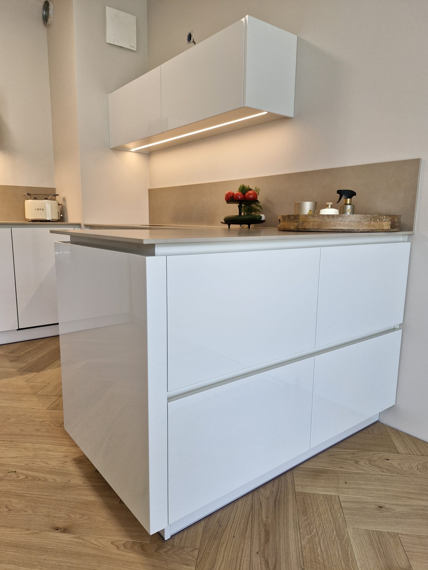 Biała, lakierowana wyspa kuchenna z szufladami i blatem, oświetlenie LED pod szafką, drewniana podłoga w jodełkę. Nowoczesne wykończenie wnętrza kuchni.