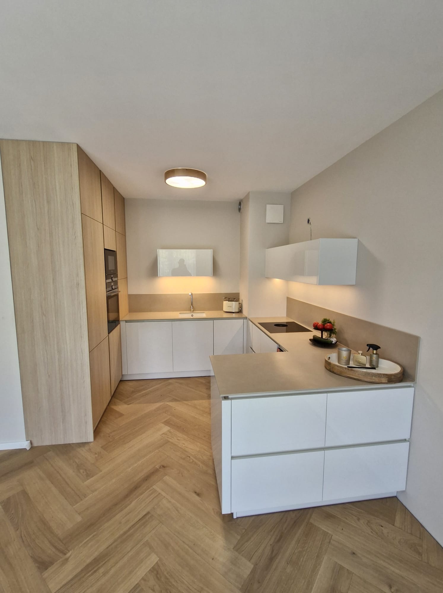 Nowoczesna kuchnia z drewnianą podłogą w jodełkę, jasnymi szafkami i blatem w odcieniach beżu. Minimalistyczne oświetlenie i dekoracje dodają elegancji.