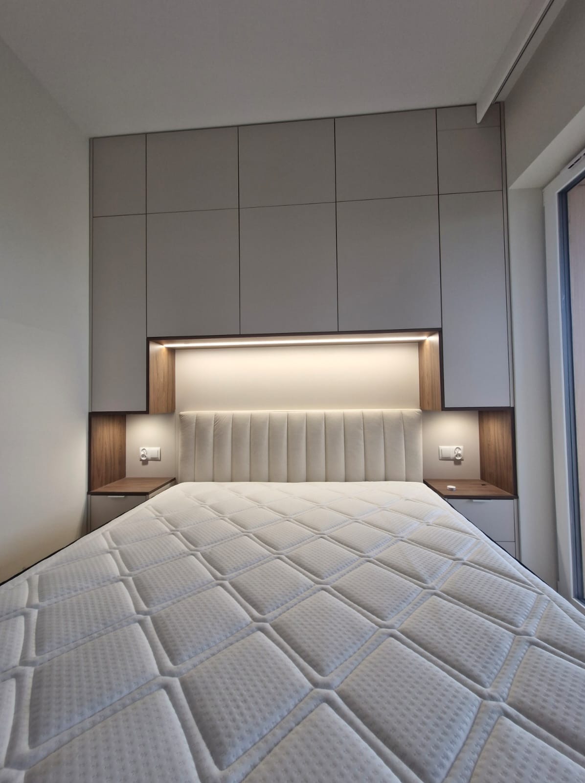 Nowoczesna sypialnia z zabudową meblową: szafy w odcieniach szarości, podświetlane półki nocne z gniazdkami i tapicerowany zagłówek łóżka. Minimalistyczny design.