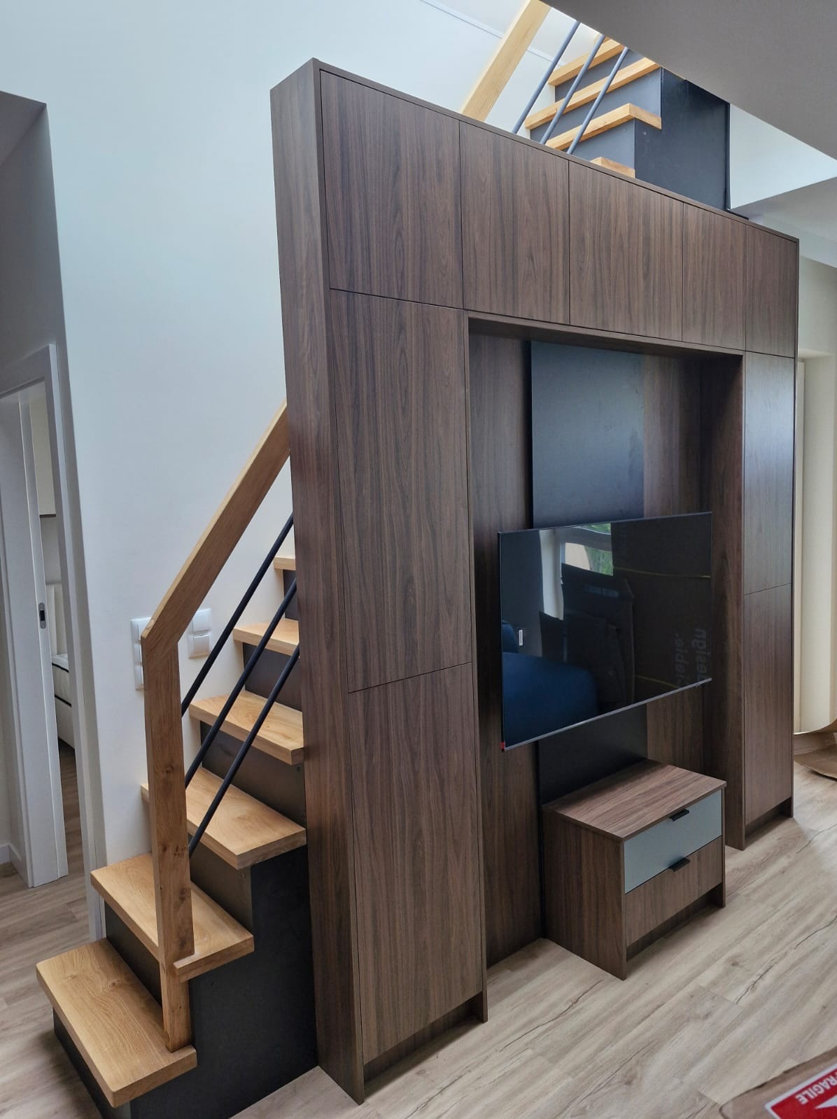 Nowoczesne schody z drewnianymi stopniami i czarnymi balustradami, wkomponowane w zabudowę meblową z ciemnego drewna, z wnęką na telewizor i szafką. Minimalistyczny design.