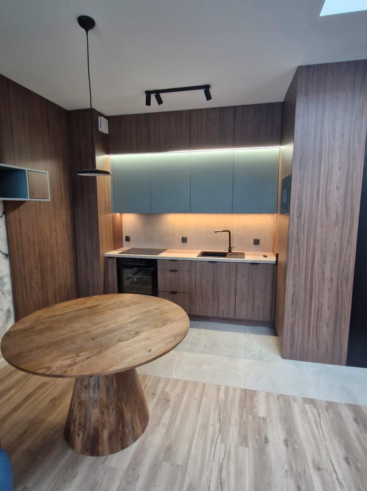 Nowoczesna kuchnia z drewnianymi frontami, oświetleniem LED pod szafkami i okrągłym stołem z drewnianym blatem na tle ściany z forniru.