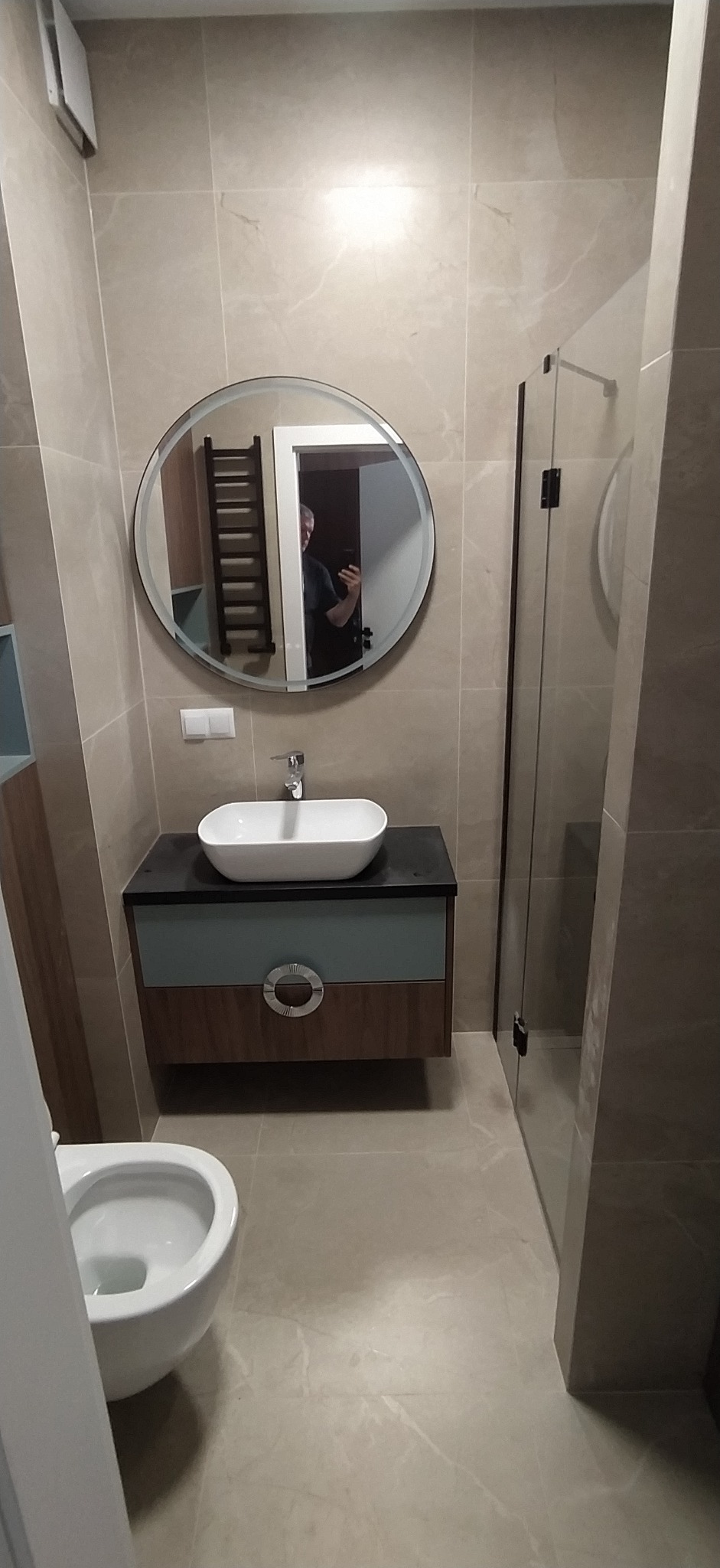 Nowoczesna łazienka z okrągłym lustrem, białą umywalką na szafce w odcieniach szarości i brązu oraz kabiną prysznicową z przezroczystego szkła.