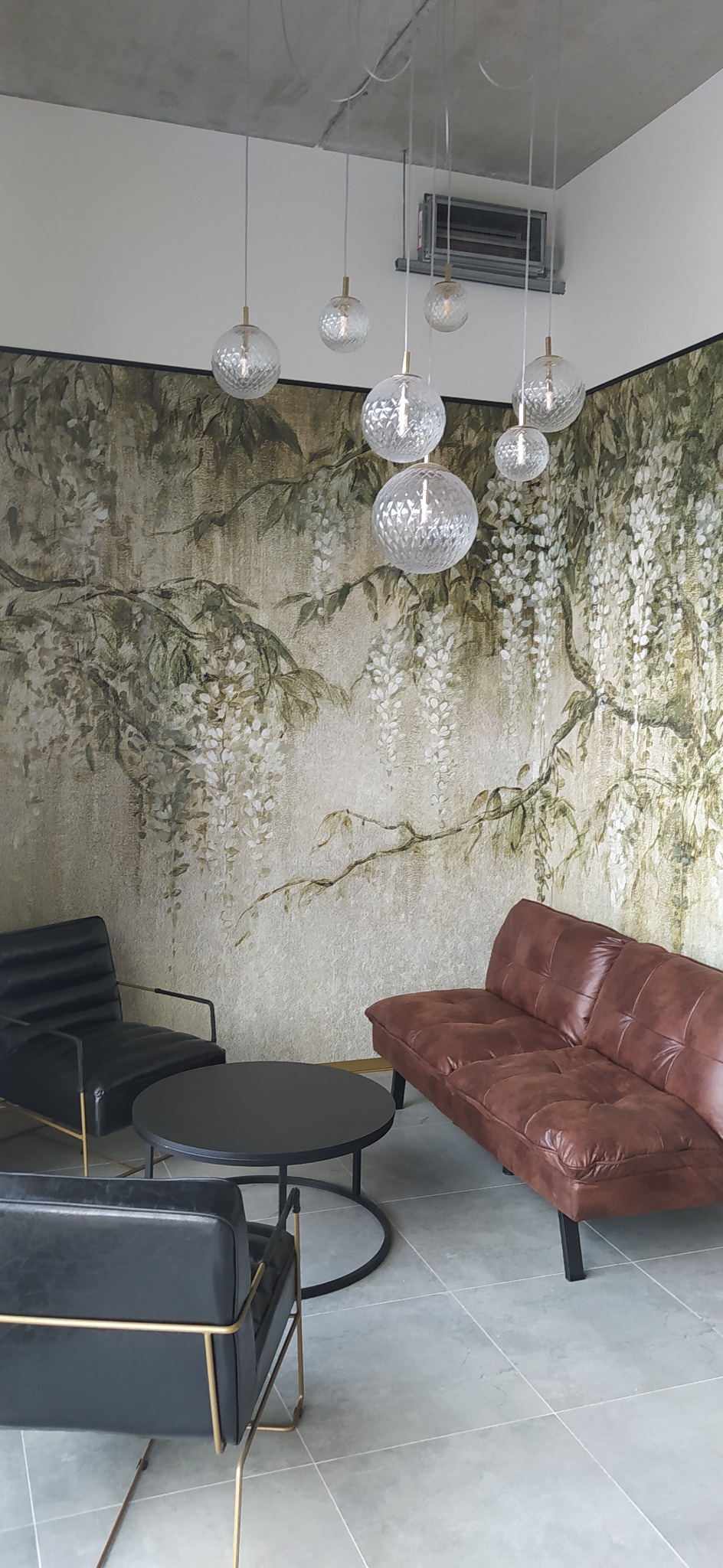 Stylowe wnętrze z tapetą w motywy roślinne, skórzaną sofą i czarnymi fotelami. Nad stolikiem wiszą szklane lampy w kształcie kul. Widoczny sufit z klimatyzacją.