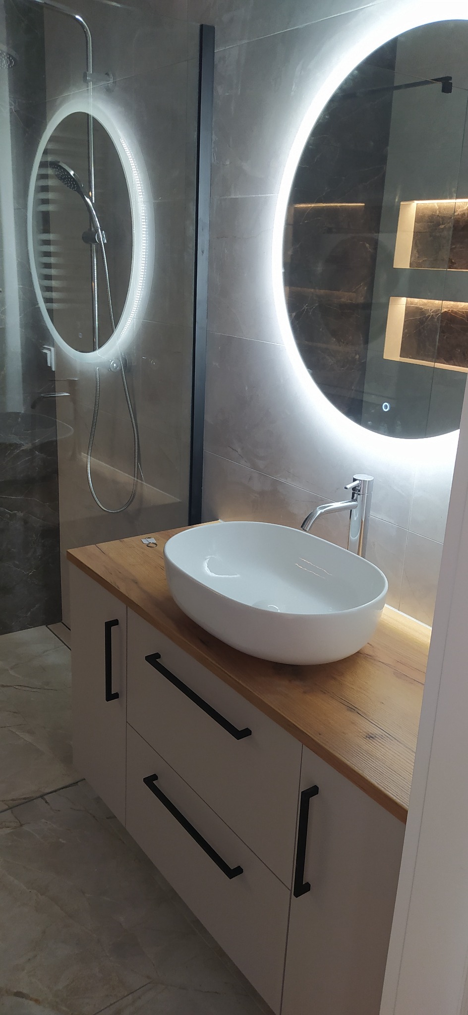 Nowoczesna łazienka z owalną umywalką na drewnianym blacie, podświetlane lustra i szklana kabina prysznicowa. Minimalistyczne, czarne uchwyty szafek.