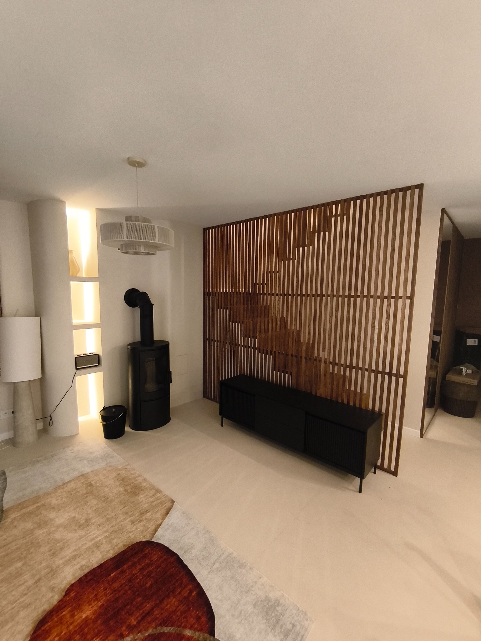 Nowoczesny salon z drewnianą ażurową ścianką działową, czarnym piecem wolnostojącym i podświetlaną wnęką. Minimalistyczny design, ciepłe oświetlenie.