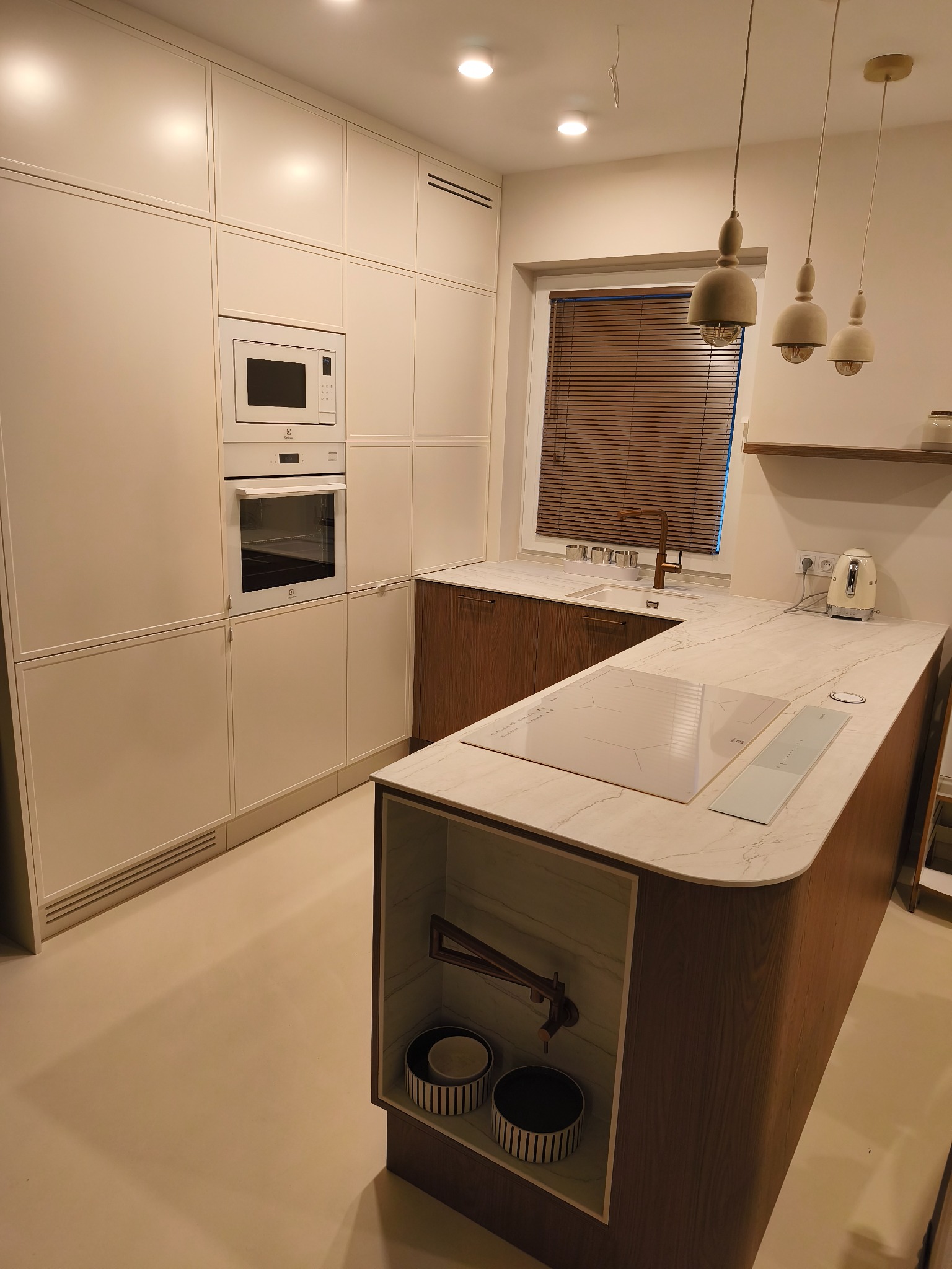 Nowoczesna kuchnia z wyspą: białe szafki, drewniane akcenty, marmurowy blat. Minimalistyczne lampy wiszące i zabudowane sprzęty AGD. Widok na okno z żaluzjami.