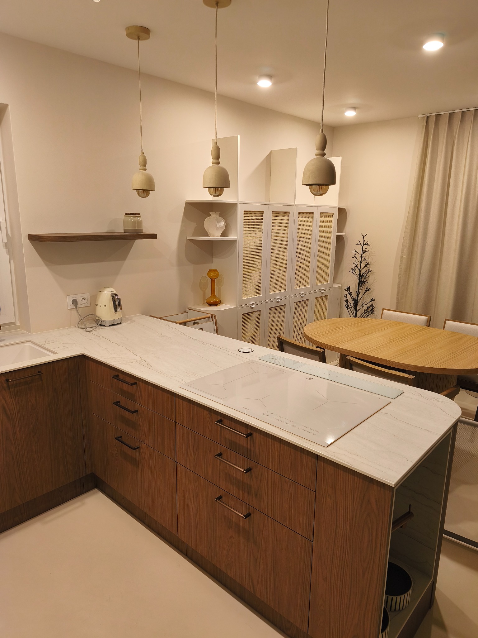 Nowoczesna kuchnia z drewnianymi szafkami, marmurowym blatem i płytą indukcyjną. Nad blatem wiszą trzy lampy w stylu skandynawskim. W tle stół i krzesła.