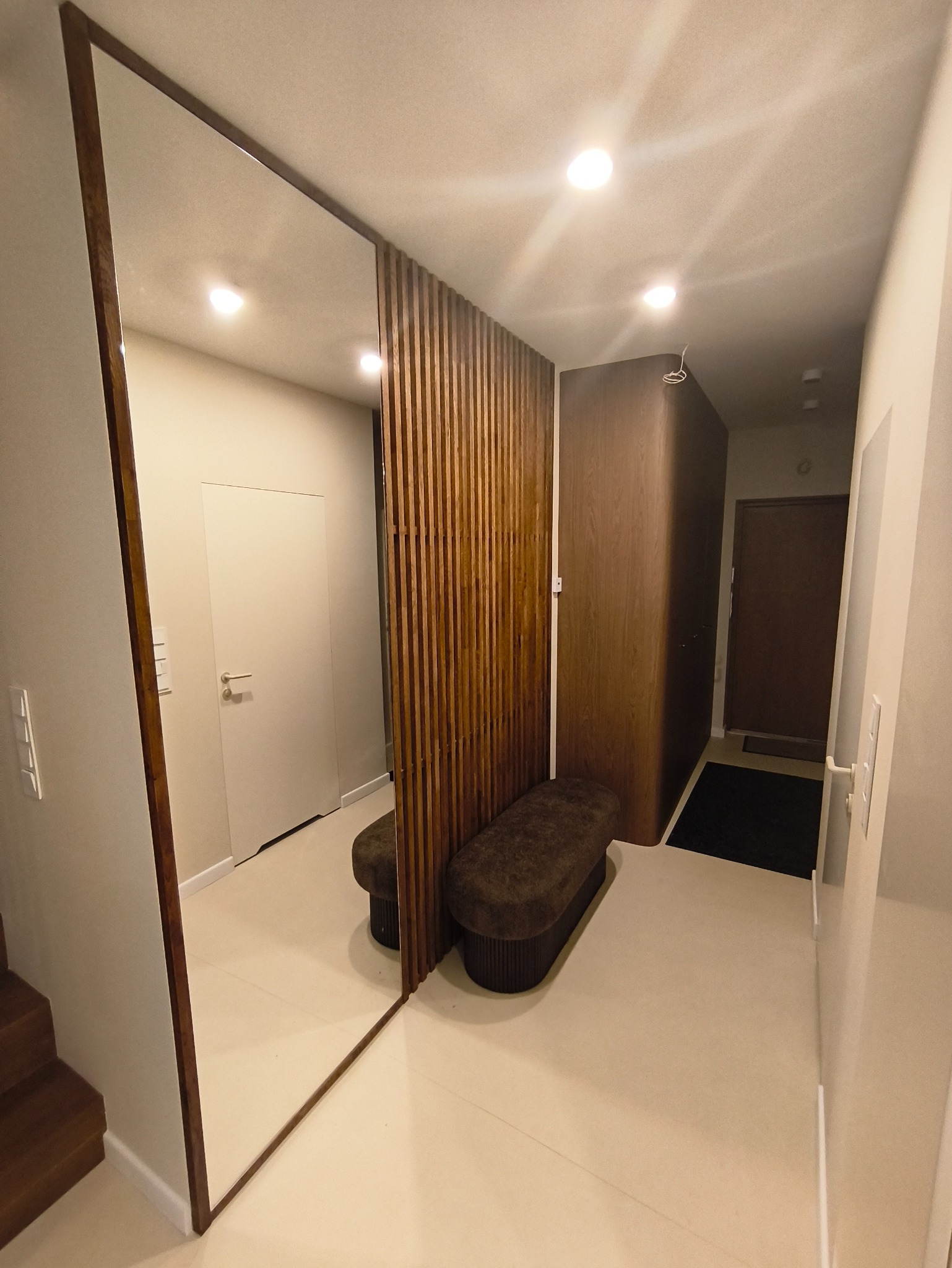 Nowoczesny przedpokój z dużym lustrem w drewnianej ramie, drewnianą ścianką działową i tapicerowaną ławką. Jasna podłoga i oświetlenie punktowe.