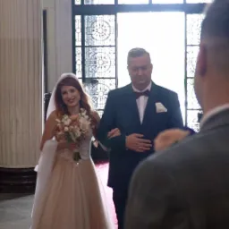 Panna młoda prowadzona do ołtarza przez ojca w kościele, widziana z perspektywy gościa w szarym garniturze, z widocznym oknem z ornamentami w tle.