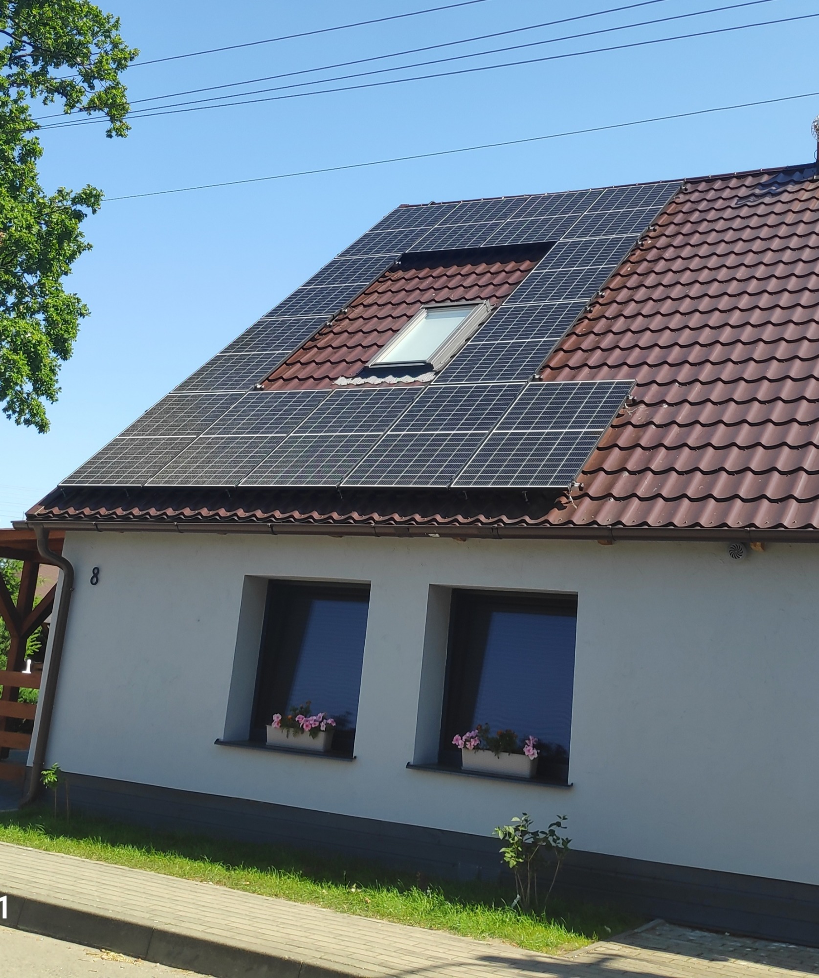 Dom jednorodzinny z brązowym dachem pokrytym panelami fotowoltaicznymi, wkomponowanymi wokół okna dachowego; widoczne dwa okna z kwiatami w doniczkach na parapecie.