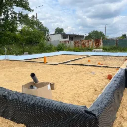 Przygotowanie fundamentów pod budowę domu: wykop z izolacją przeciwwilgociową, piasek zasypowy, widoczne rury kanalizacyjne w wykopie, w tle budynek gospodarczy i zieleń.