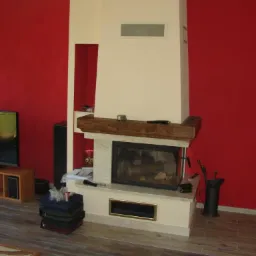 Wykończony kominek z drewnianą półką w salonie z czerwoną ścianą w Tczewie. Beżowa obudowa kominka z jasnymi elementami dekoracyjnymi i paleniskiem.