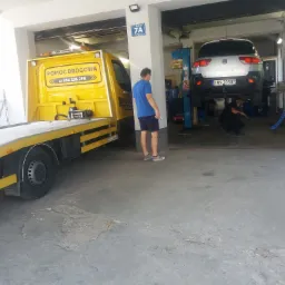 Wnętrze warsztatu samochodowego z podnośnikiem, na którym stoi srebrny samochód z widoczną tablicą rejestracyjną, oraz żółtą lawetą pomocy drogowej na zewnątrz. Pracownik warsztatu pracuje...