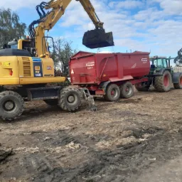 Żółta koparka kołowa Komatsu PW148 ładuje ziemię do czerwonej przyczepy ciągniętej przez zielony traktor Fendt na placu budowy, z widoczną niwelacją terenu i innymi maszynami rolniczymi w tle...