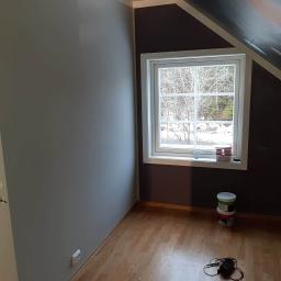 Lukmar - Pokój na poddaszu po malowaniu: ściana w odcieniach szarości, brązowa ściana przy oknie z widokiem na zimowy krajobraz, drewniana podłoga, puszki po farbie i miernik elektryczny na podłodze.