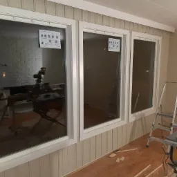 Trzy okna z białymi ramami zamontowane w ścianie z boazerią, widoczne narzędzia i drabina wskazują na trwający montaż.