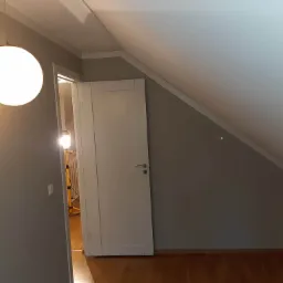 Pokój na poddaszu w trakcie odświeżania, ściany pomalowane na szaro, widoczna biała lampa kula, otwarte białe drzwi i lampa warsztatowa w tle.