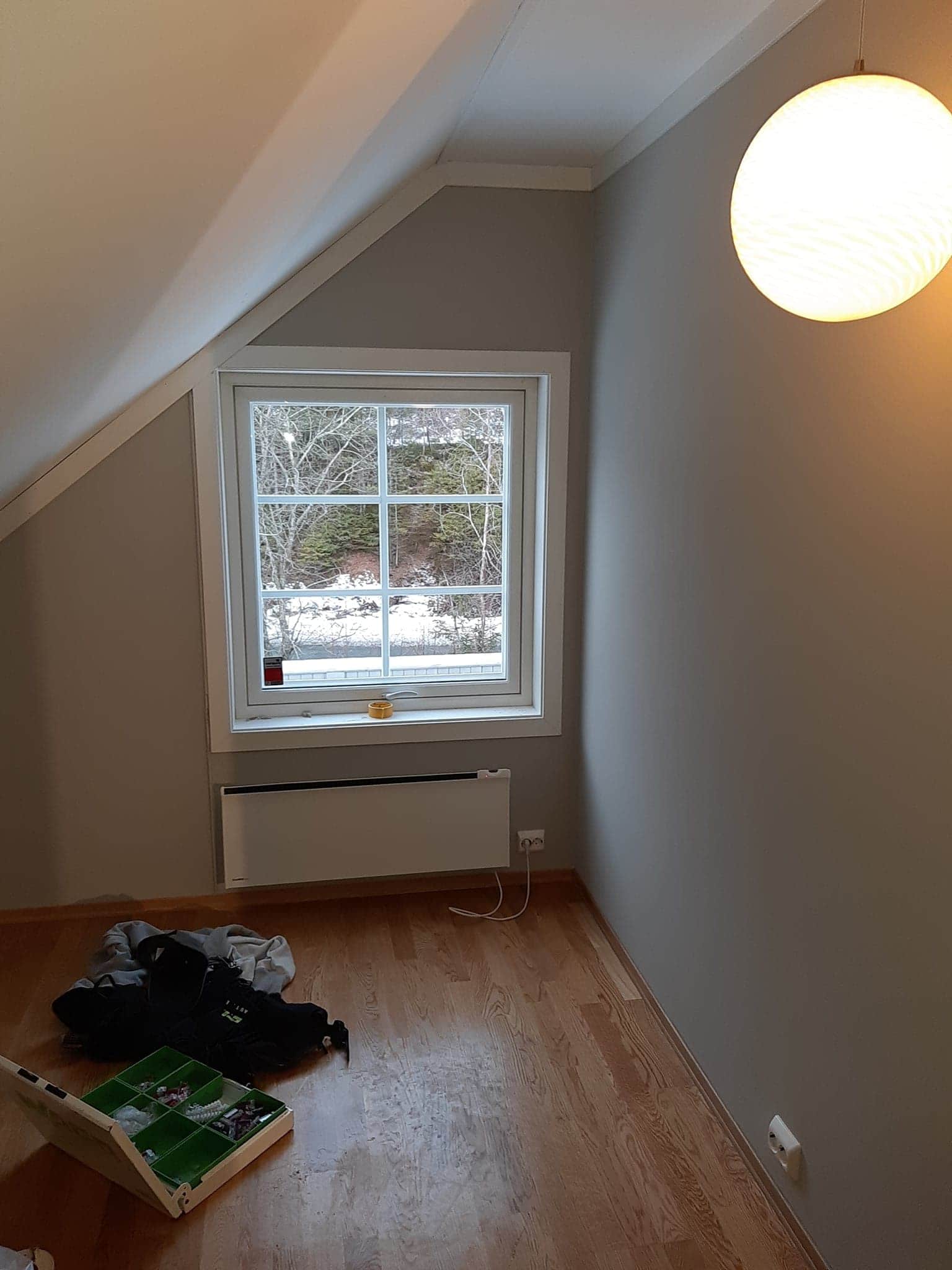 Pokój na poddaszu z oknem wychodzącym na zimowy krajobraz, drewniana podłoga, szare ściany, widoczna lampa sufitowa oraz pudełko z biżuterią i ubrania na podłodze.