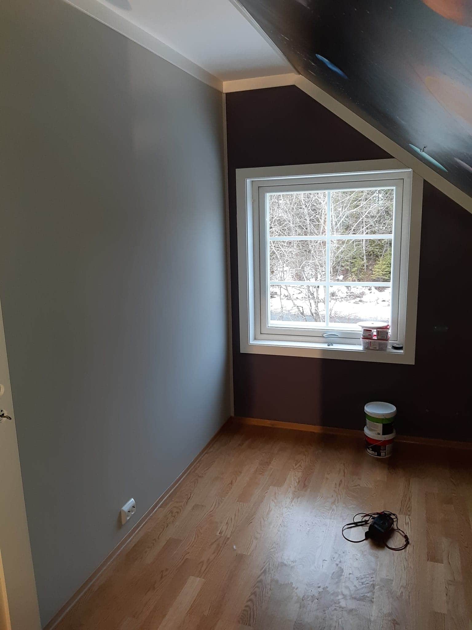 Pokój na poddaszu po malowaniu: ściana w odcieniach szarości, brązowa ściana przy oknie z widokiem na zimowy krajobraz, drewniana podłoga, puszki po farbie i miernik elektryczny na podłodze.