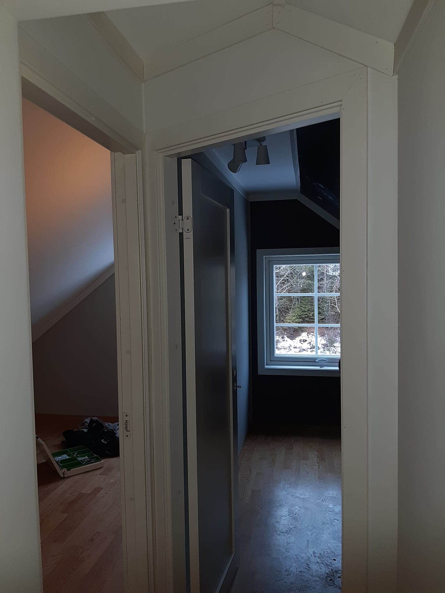 Widok na pokój na poddaszu przez otwarte drzwi z białą ościeżnicą. W pokoju widoczne okno z widokiem na las zimą i drewniana podłoga. W tle, w drugim pokoju, widoczna podłoga i częściowo widoczny...