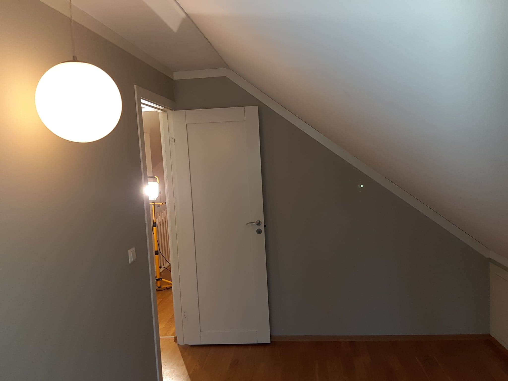 Pokój na poddaszu w trakcie odświeżania, ściany pomalowane na szaro, widoczna biała lampa kula, otwarte białe drzwi i lampa warsztatowa w tle.