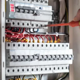 Szczegółowe ujęcie tablicy rozdzielczej z bezpiecznikami, podczas podłączania przewodów przez elektryka za pomocą pomarańczowego śrubokręta.