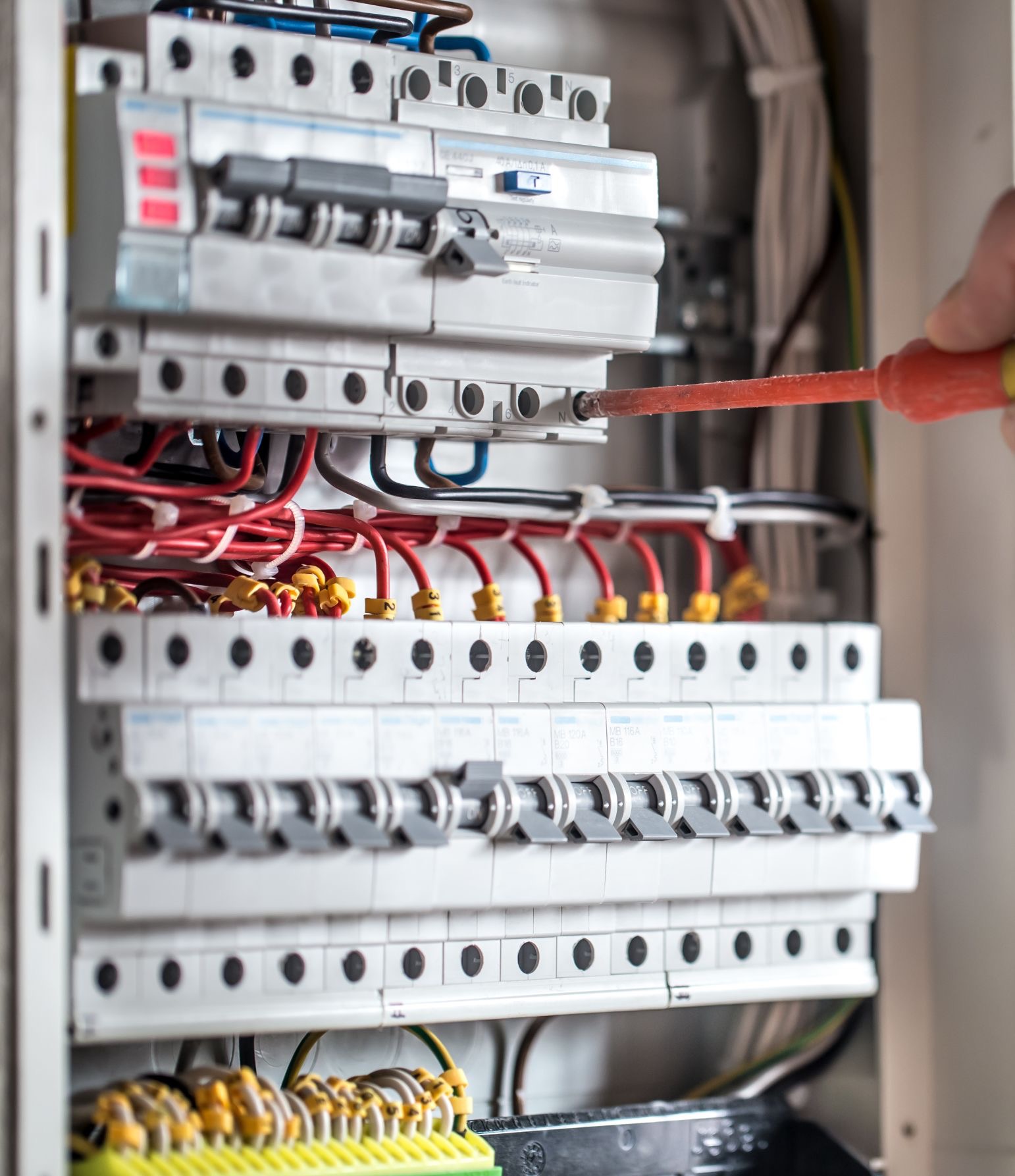 Szczegółowe ujęcie tablicy rozdzielczej z bezpiecznikami, podczas podłączania przewodów przez elektryka za pomocą pomarańczowego śrubokręta.